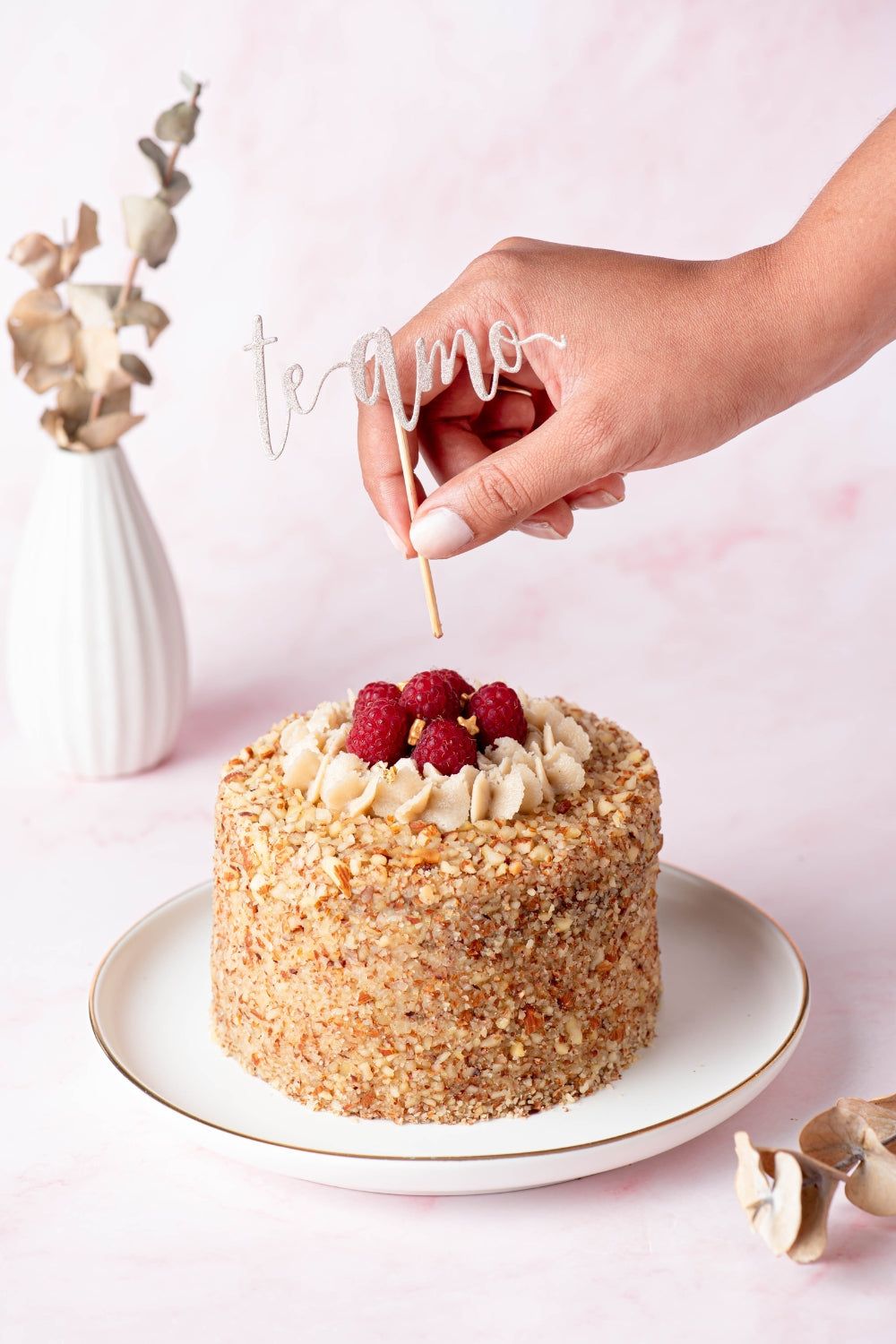 Mini torta de zanahoria en un plato color blanco sobre un fondo rosado pastel. Una mano se acerca al postre para insertar un topper con las palabras "te amo" en el postre. 